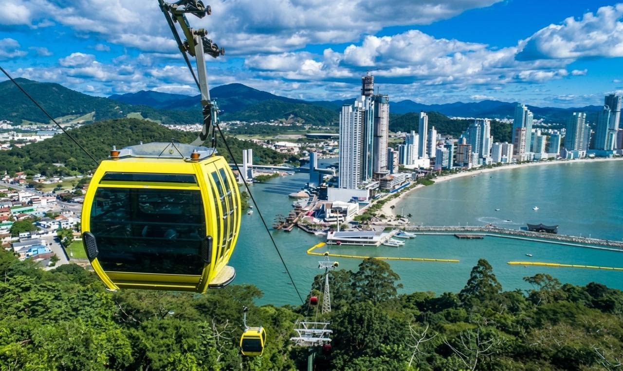 Bondinho amarelo sobre floresta verde com vista para arranha-céus e praia de Balneário Camboriú.